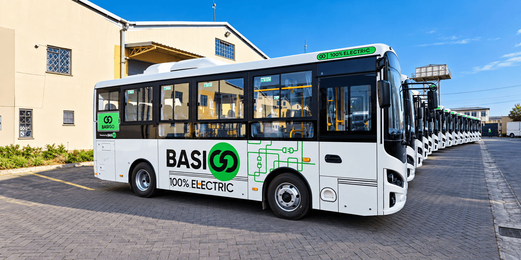 A row of white BasiGo electric buses parked outside an assembly facility.