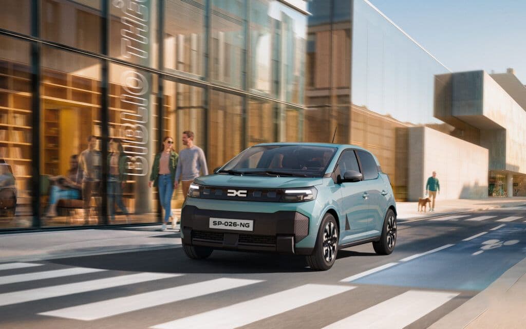 A light blue Dacia Spring electric car driving through a European city.