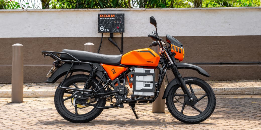 Orange Roam Air electric motorcycle fitted with a Gen-3 battery beside a Roam fast charger